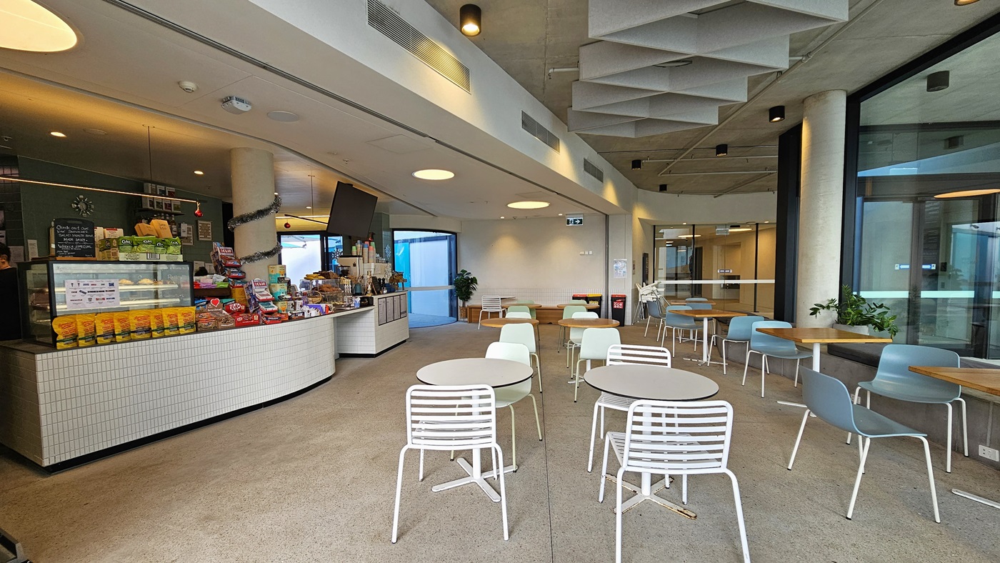 The cafe's reception counter features small white tiles. Food racks, snacks, and treats are displayed on the countertop. The left section holds four posters with the menu, drinks dispenser, and plastic cups. A display rack showcases light snacks on the right. A flat-screen TV is ceiling-mounted. Christmas decorations adorn the space. The ceiling has diamond-shaped wooden structures, and the right wall is glass. Movable tables and chairs are positioned on the right.