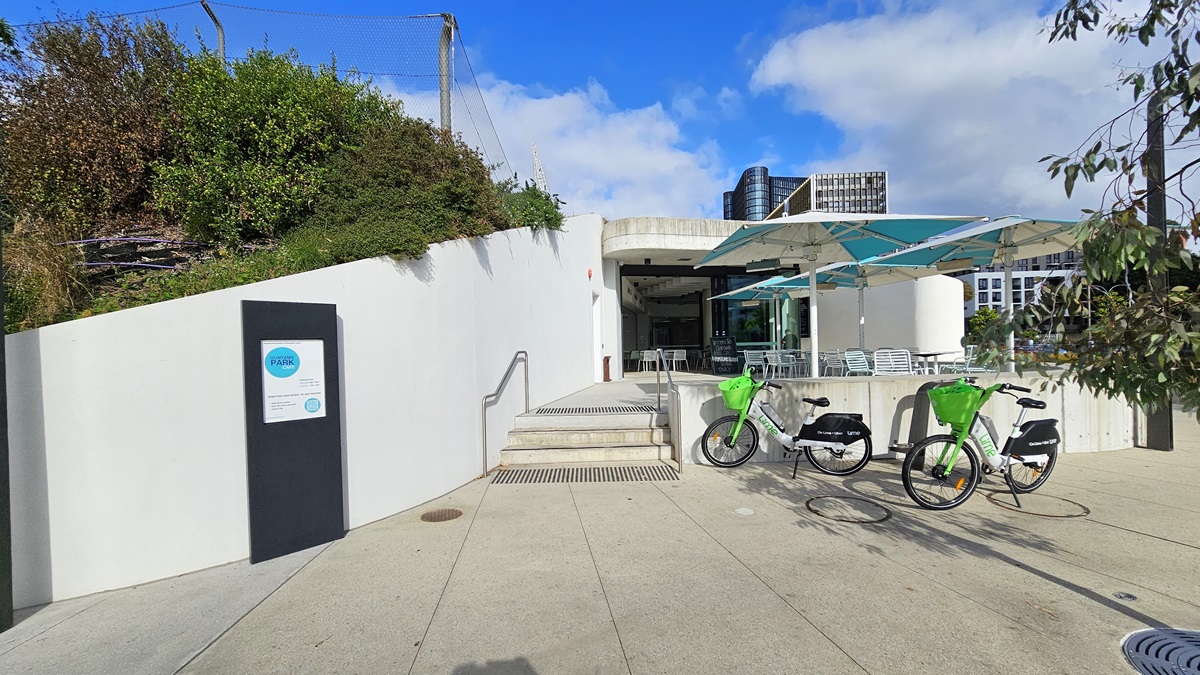 To the left is a stepped entry equipped with handrails, preceded by truncated domes on the floor. A signage displaying the facility name is positioned on the white concrete wall near a hillside with shrubs. Upstairs is the cafe, featuring large umbrella shades and arranged tables and chairs. Two bikes are parked in front of the facility, near a tree. A tall commercial building is visible behind the structure.
