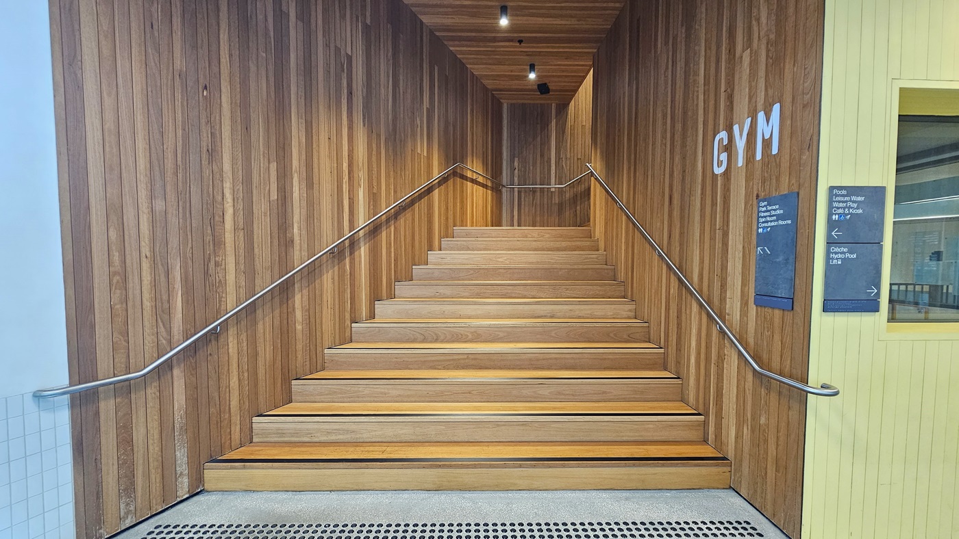 Access to the gym is through a stairway with wooden panels, equipped with handrails and truncated domes. A prominent sign reading "GYM" is positioned on the right wall. The walls and ceiling feature a timber design with LED lights. A signage is posted on the right wall.