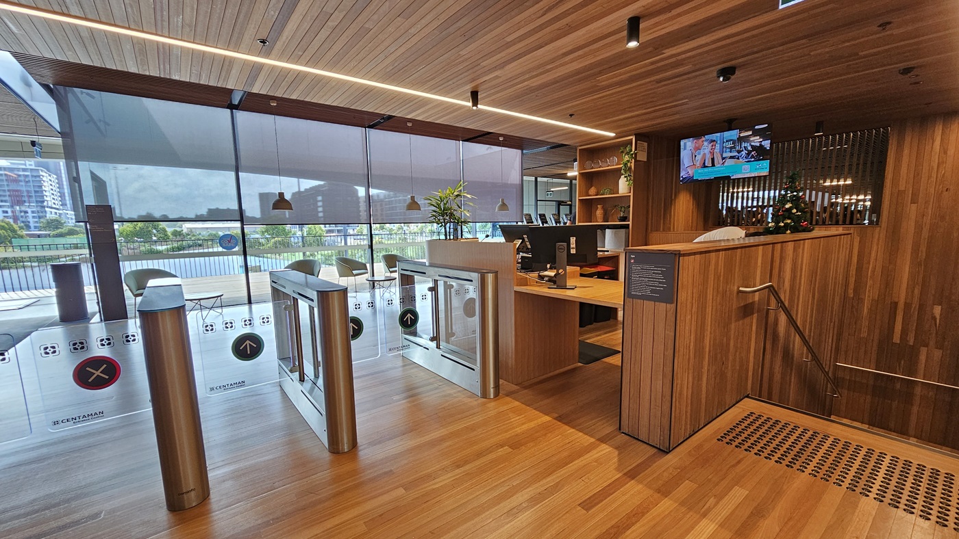 At the top of the stairs is the entrance to the gym, with handrails and truncated domes on the stairs. To the left is a gated entry with glass turnstiles marked for exit and entry. The reception desk on the right features a timber design. A flat-screen TV is mounted on the ceiling. Inside, there are movable plastic chairs and tables, and a retractable blind covers the glass wall. The ceiling, floor, and walls all feature timber designs.
