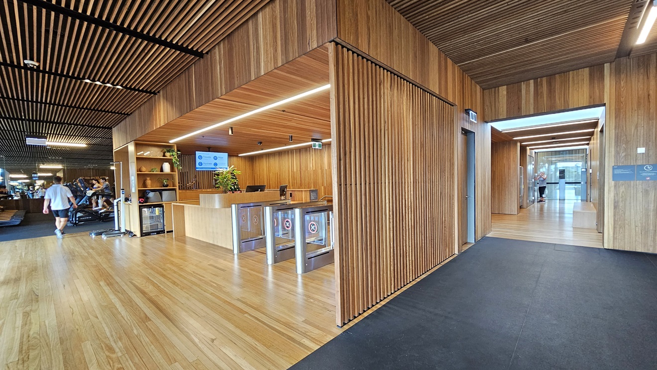 On the left is a gated entry with glass turnstiles marked for exit and entry. The reception desk on the right features a timber design. A flatscreen TV is mounted on the ceiling. A display rack with jar decorations is present. The ceiling, floor, and walls all feature timber designs. To the left of the reception is the gym, while the lobby on the right leads to a corridor. The signage is affixed to the right wall.