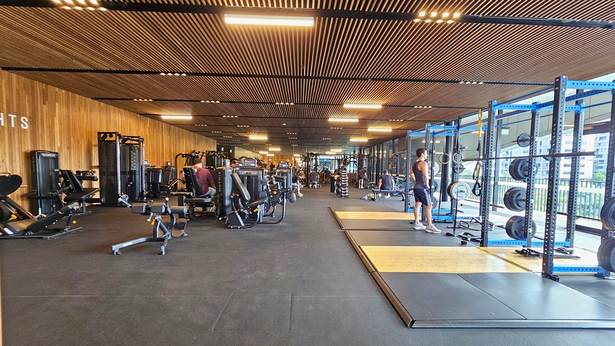 To the right is a glass wall providing an outside view. Smith machines are positioned nearby. On the left is the weights area with bench press and inclined machines lined up, and more machines displayed further in. The floor features black rubber tiles with a slightly elevated section on the right. The ceiling has wooden plank designs with fluorescent lights. Gym users are visible.