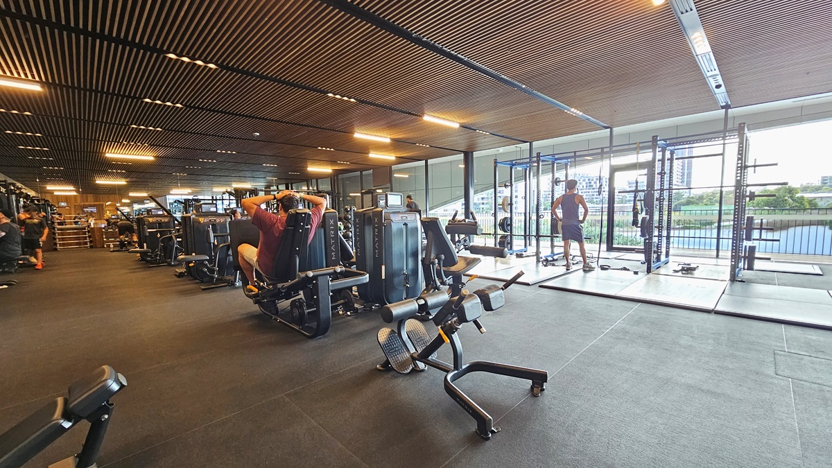 To the right is a glass wall providing an outside view. Smith machines are positioned nearby. In the middle are additional resistance machines. On the left, bench press machines and other equipment are displayed further in. The floor features black rubber tiles with a slightly elevated section on the right. The ceiling has wooden plank designs with fluorescent lights. Gym users are visible.