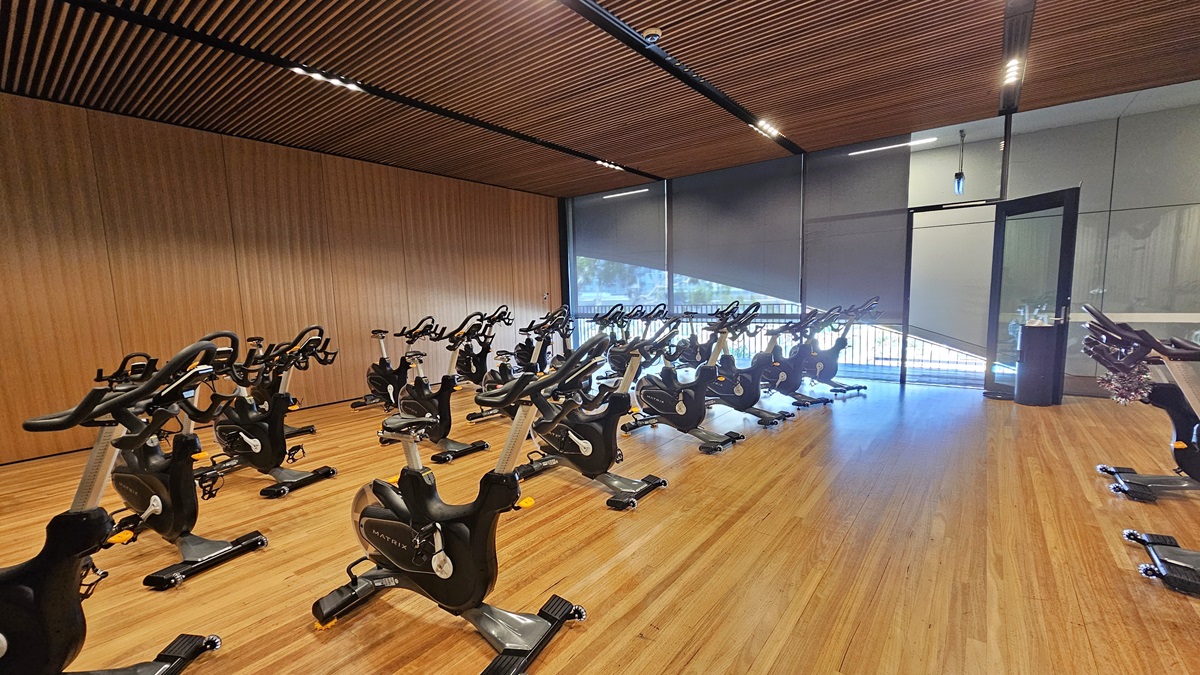 Spin machines are arranged in rows, filling the timber-floored room. An inward-opening glass door serves as an entry to this space. A black rubbish bin is placed near the door. The right wall is made of glass, and the left wall has a timber design with a wooden plank ceiling design equipped with LED lights.
