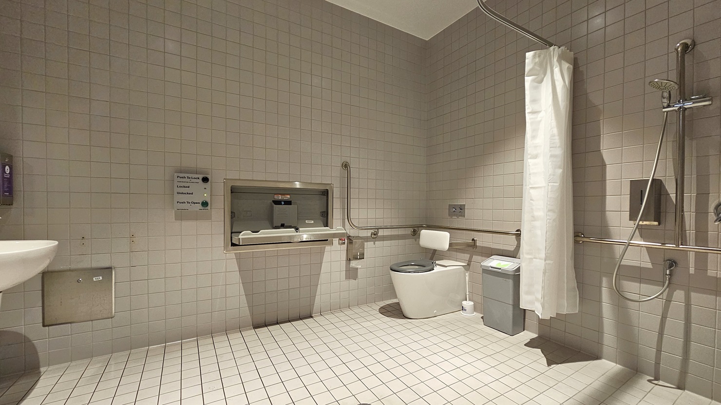 Inside the accessible bathroom, a toilet bowl equipped with handrails is positioned in the left corner. Beside it is a rubbish bin, toilet brush, and tissue dispenser. Adjacent to it is a shower with a curtain and hand grabs. In the middle is a change table with an access button. A washbasin and a soap dispenser are placed on the left wall. The walls are covered with white tiles from floor to ceiling.