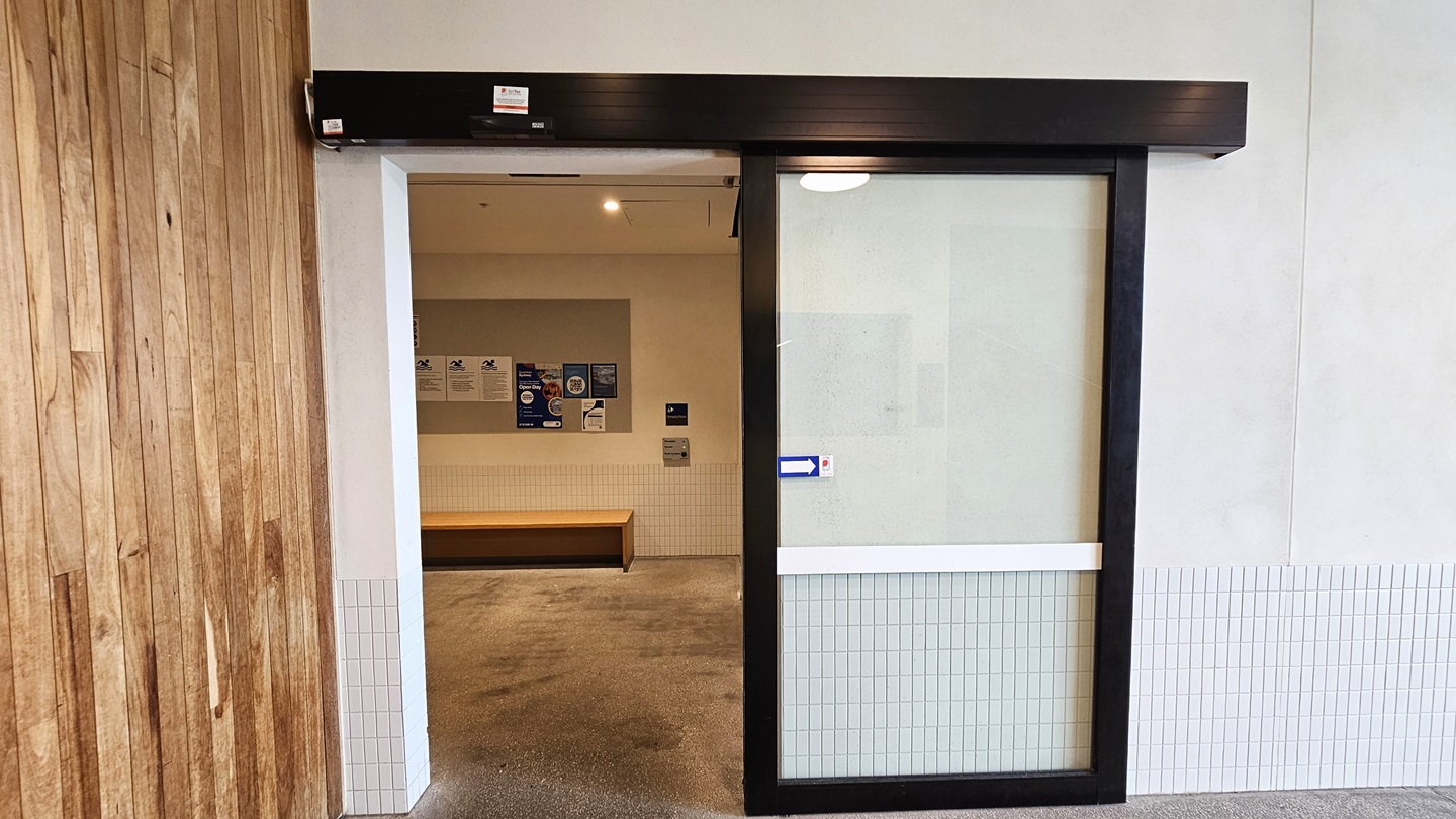 The entrance to the Hydrotherapy Pool is through an automatic glass sliding door. The left wall features a timber design, and the right wall is white concrete. Inside, the wall is equipped with safety banners and reminder signs. A wooden bench chair is placed against the wall.