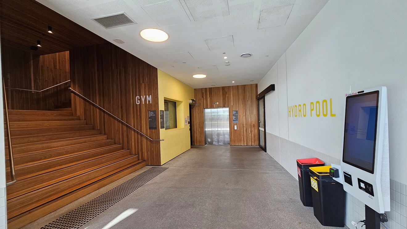 To the left are wooden stairs equipped with handrails and truncated domes. On the right is a door leading to the Hydro Pool. The left walls and doors have a timber design. A self-service kiosk is positioned on the right. The floor on the right is white, bearing the "Hydro Pool" sign. The ceiling features white with LED lights.