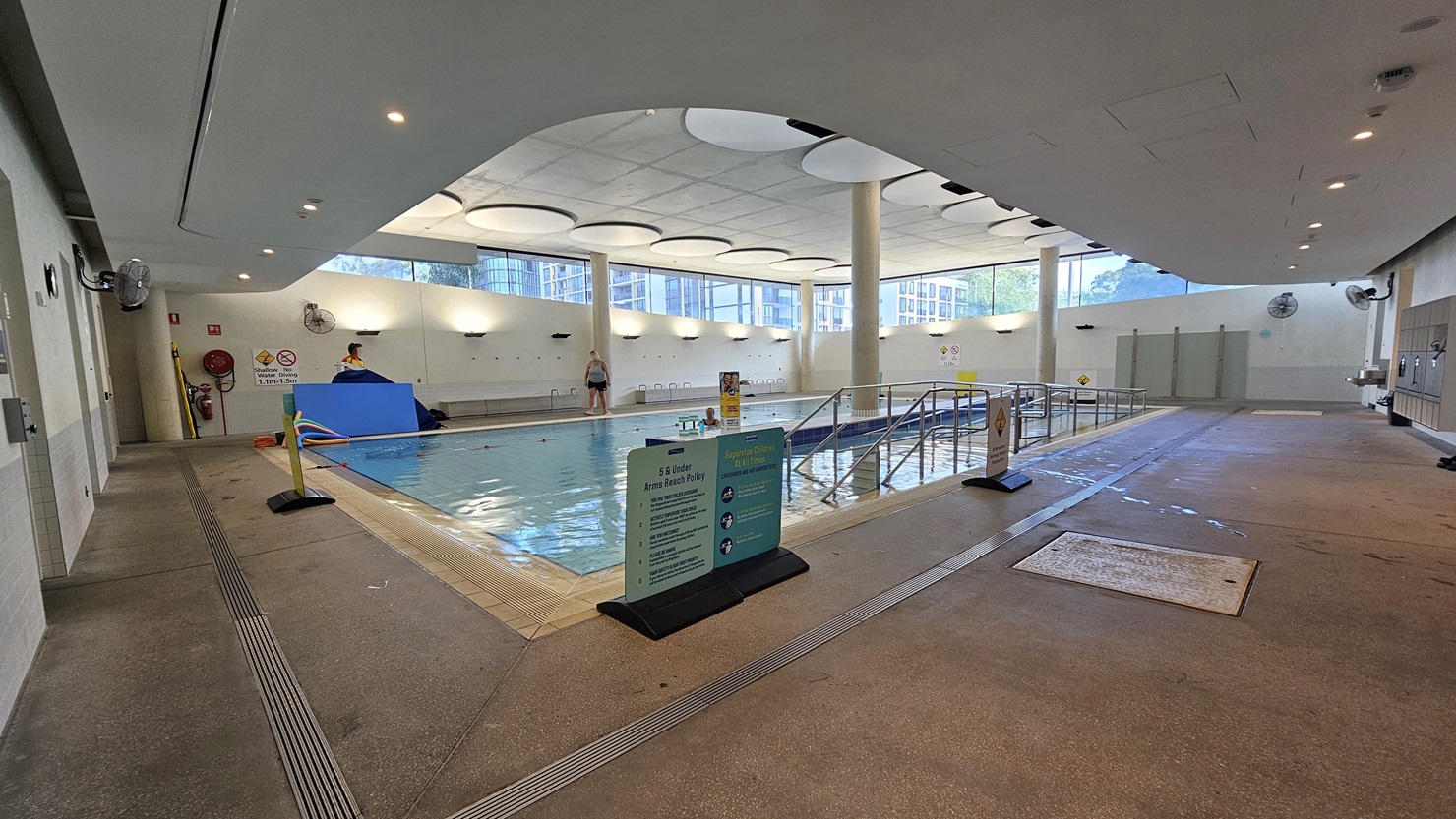 The Hydrotherapy Pool features a step and ramp entry with handrails. Free Standing banners are placed on the pool deck, and the pool is surrounded by an overflow grid. On the left wall, a wall fan, fire hose reel, and fire extinguisher are mounted. Floating beds and hoses are also visible. The upper wall is made of glass, and the ceiling has circular-shaped decorations.