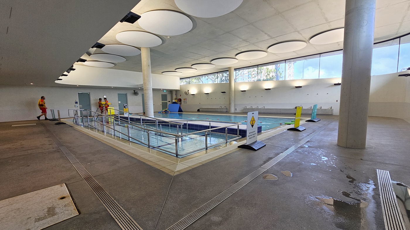 The Hydrotherapy Pool is designed with step and ramp entries, each equipped with handrails. Freestanding banners adorn the pool deck, and the pool is encircled by an overflow grid. Floating beds and noodle hoses are visible. The upper wall is made of glass, providing an outside view, and the ceiling is adorned with circular-shaped decorations and floodlights. Lifeguards are present.