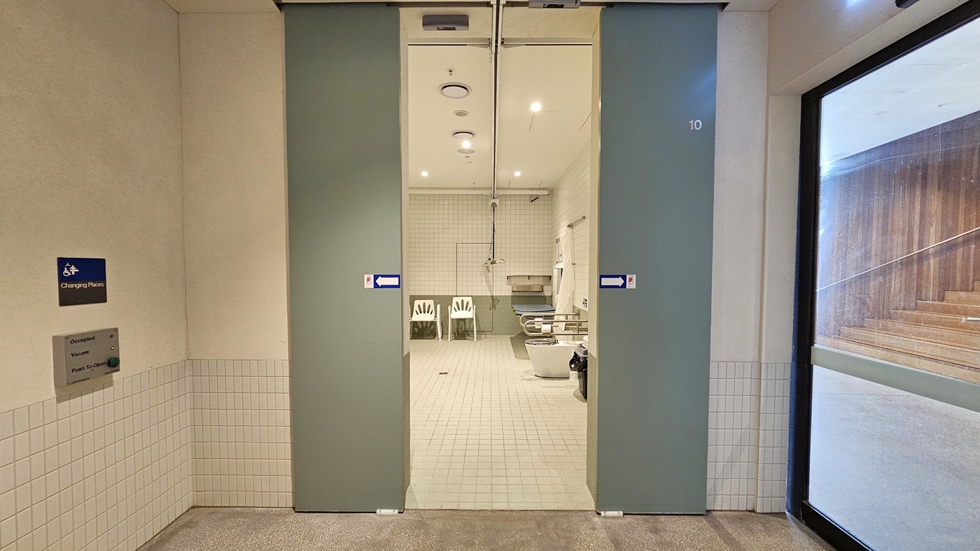 The accessible change room features sliding doors with signage and access buttons on the left wall. Inside, the toilet bowl with hand grabs, two white plastic chairs, and a change table are visible. The ceiling is all white, and the walls are white with green accents. To the right, the wooden stairs are visible through the glass wall.