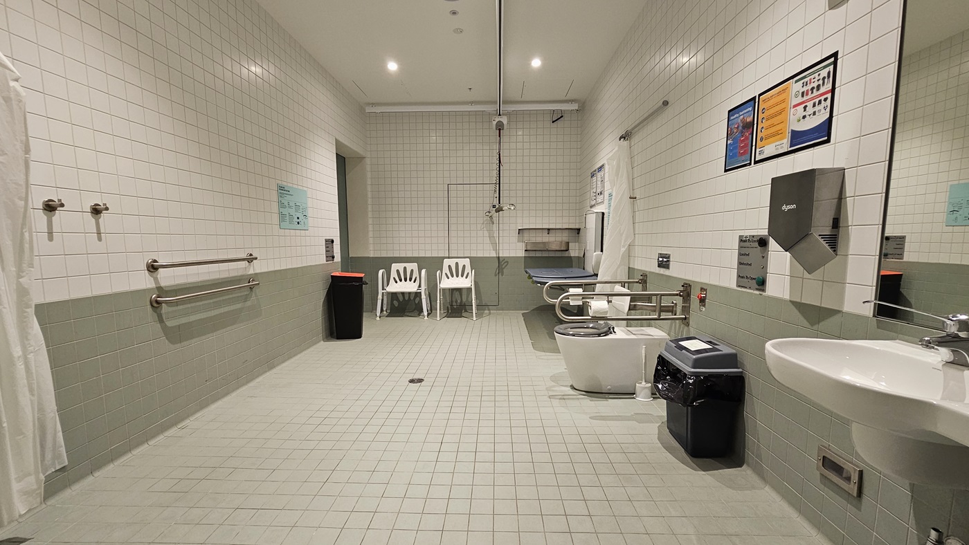 On the right, there's a foldable change table with a ceiling hoist. Adjacent is the toilet bowl with hand grabs, rubbish bins, and access buttons. A wash basin, mirror, and another rubbish bin are on the right. The ceiling is all white, and the walls are white with green accents. To the right, the wooden stairs are visible through the glass wall.