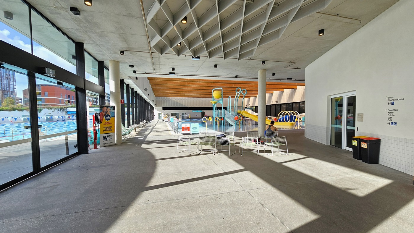 In the middle, movable white chairs with mesh designs are arranged. To the right, a white wall with signage for the accessible toilet is present. Rubbish bins are visible. Behind, the Aqua Play Pool features a yellow slide with fountains, tipping buckets, and water features. A retractable banner is positioned near the glass wall, and safety banners are placed on the pool deck. The ceiling is white with diamond-shaped wooden structures and wooden planks.