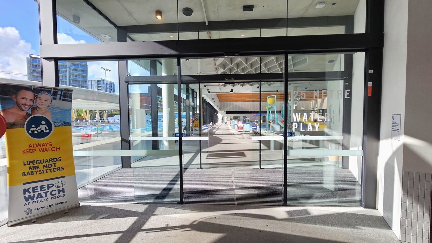 The entrance to the Aquatic facility is through double automatic glass doors equipped with non-slip mats. A large retractable banner is positioned on the left. Inside the indoor facility, the Aqua Play Pool is visible first on the right. The ceiling features white with diamond-shaped wooden structures and LED lights.
