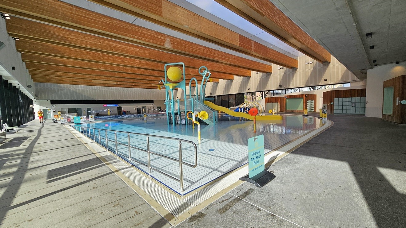 The Aqua Play Pool includes a beach entry with a yellow slide, fountains, tipping buckets, and water features. A ramp entry with handrails extends to the other side of the pool on the left. The pool is surrounded by an overflow grid. The ceiling features a wooden plank design with floodlights. To the far left, there is an outdoor shower and Cabana-design bathrooms. Safety banners are visible on the pool deck.