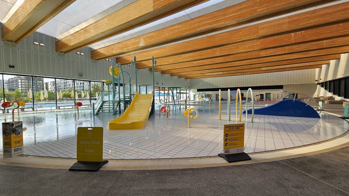 The Aqua Play Pool has colourful structures, a yellow slide with sprayers, tipping buckets, fountains, and other water features, including a wading area. An overflow grid surrounds the pool, and safety banners are placed on the pool deck. The ceiling features a wooden plank design with floodlights. The left wall has glass walls providing a view of the outdoor aquatic facility.