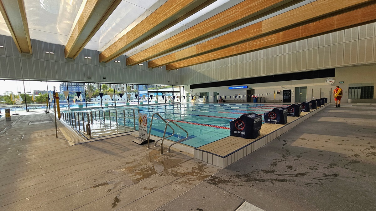 The pool features a ramp entry with a handrail. Diving boards with covers are arranged on the raised pool deck on the right, with safety signs placed on the pool deck. To the far right, accessible bathrooms and outdoor showers are present. A round clock is affixed to the upper wall. On the far end, a glass wall provides a view of the Aquatic Facility. The ceiling features transparent roofing with wooden plank accents.