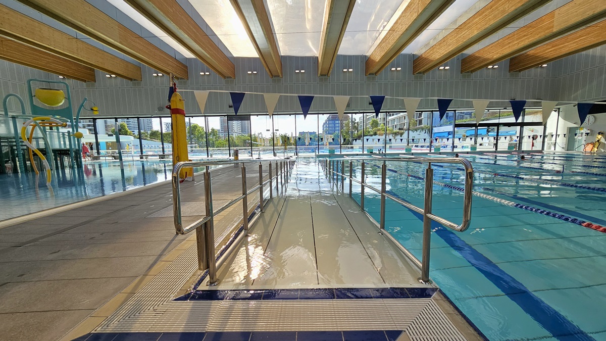 To the left is the Aqua Play Pool equipped with slides, tipping buckets, and water features. Adjacent to it is the Program Pool featuring a ramp entry with handrails, floating ropes, and blue and white triangle banners. In between the two pools is a pool deck that serves as a pathway. An overflow grid surrounds both pools, and a hoist is positioned nearby. The middle wall has glass providing a view of the outdoor aquatic facility. The ceiling features transparent roofing with timber accents.