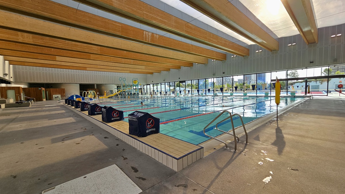 The pool is equipped with a ramp entry featuring a handrail. Diving boards with covers are arranged on the raised pool deck in the middle. To the far left, accessible bathrooms and outdoor showers are present. A round clock is affixed to the upper wall. On the far right, a glass wall provides a view of the Aquatic Facility. The ceiling features transparent roofing with wooden plank accents.