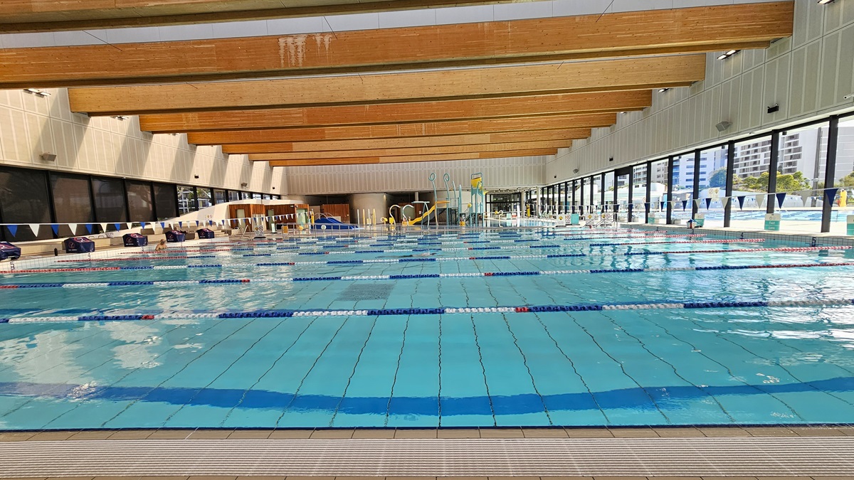 The pool is equipped with floating ropes and a string of triangle banners running across it. Diving boards with covers are lined up on the raised pool deck on the left. To the far left, accessible bathrooms and outdoor showers are present. On the right, a glass wall provides a view of the Aquatic Facility. The ceiling features transparent roofing with wooden plank accents.