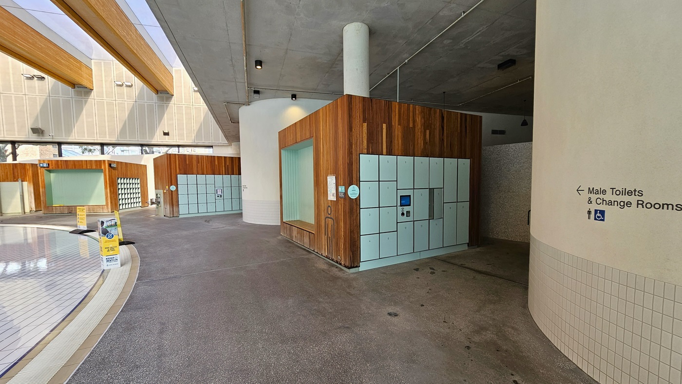 To the right are Male and Female Toilets and Change Rooms in Cabana style. The bathroom features a dark brown wood panel and white tiles design. To the left is the Aqua Play Pool, equipped with an overflow grid and safety banners. The wall on the right is white with small brick accents. A sign stating "Accessible Bathroom" is written on the wall. The ceiling features a transparent white ceiling with timber accents.