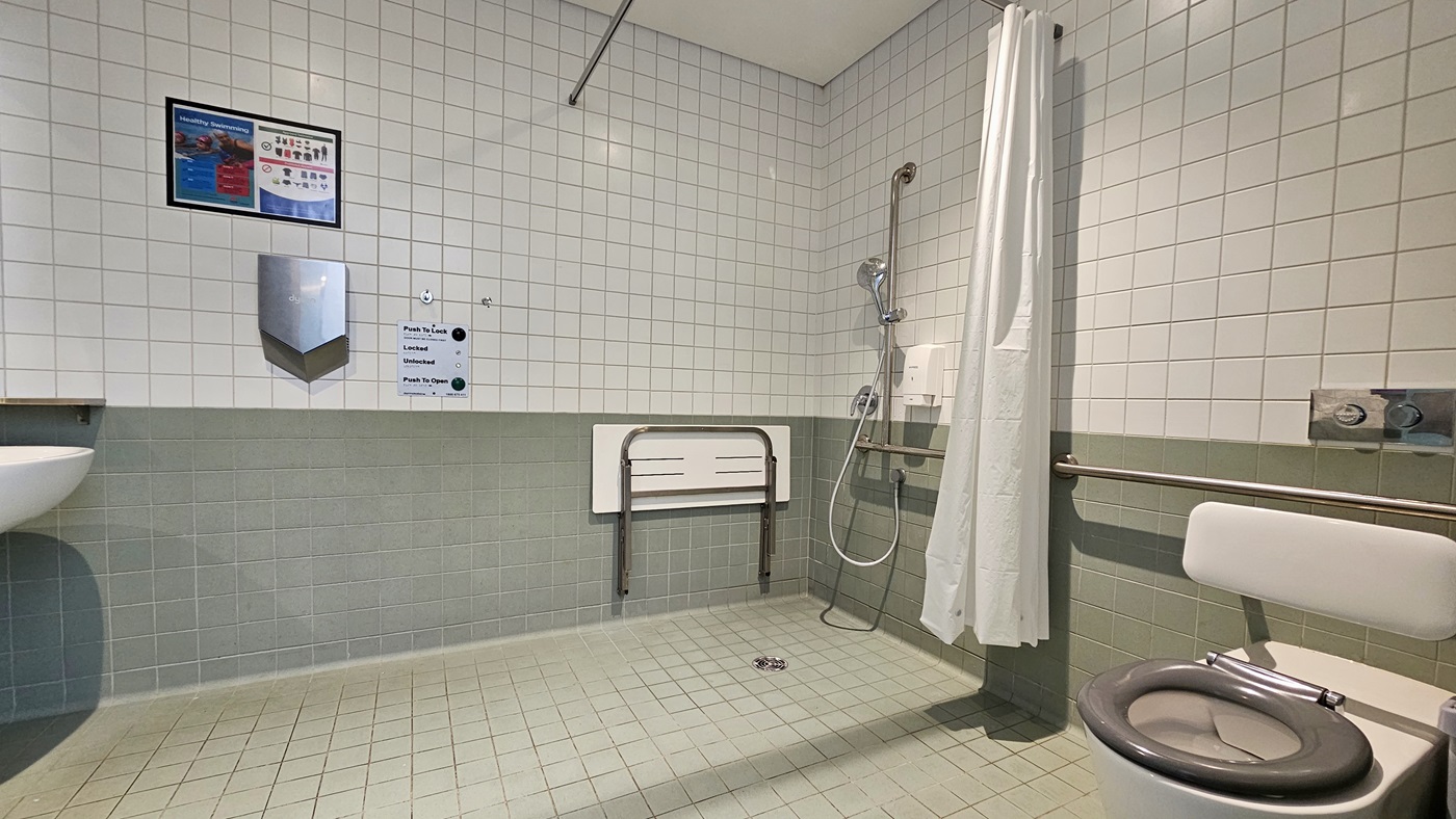 To the right is a toilet bowl equipped with handrails. Adjacent to it is a shower with a handrail and white curtains. A foldable bench chair is affixed to the wall. To the left is a wash basin and tissue dispenser. The wall is a combination of white and light green tiles, and the ceiling is all white. The floor is grey, and a poster is affixed to the wall on the right.