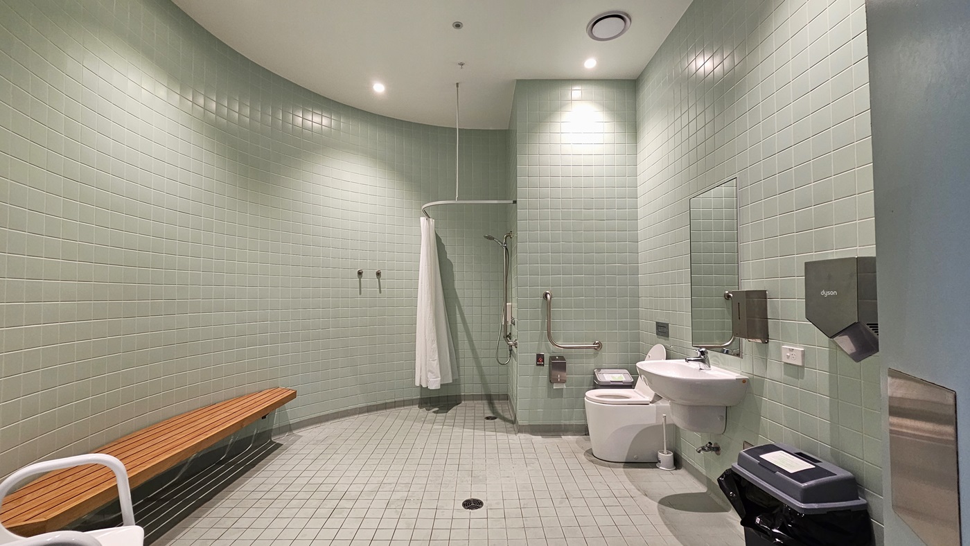 In the middle is a shower equipped with handrails and a shower curtain. Next to it is the toilet bowl with hand grabs. To the right, a washbasin, mirror, tissue, and soap dispenser are affixed to the wall. A rubbish bin and toilet brush are placed nearby. To the left is a wooden bench for seating. The walls are covered with light green tiles from floor to ceiling. The ceiling is white with LED lights, and the floor has white tiles.