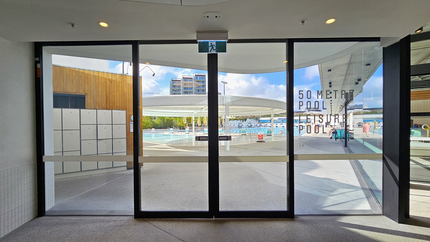 Entrance to the Outdoor Aquatic Centre is through a floor-to-ceiling automatic glass door, with an exit sign placed on top. Outside, lockers are on the left near the accessible bathroom. Immediately ahead is the Leisure Pool with a large circular shade structure.
