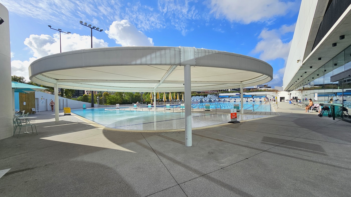 In the middle is the Leisure Pool with a large circular shade structure. Movable chairs and tables are on the left, along with safety banners. To the right, a section of the indoor facility's glass wall is visible, with bench chairs lined up against it. Further ahead is the 50m Lap Pool, with tall floodlights and folded yellow umbrella shades also visible.