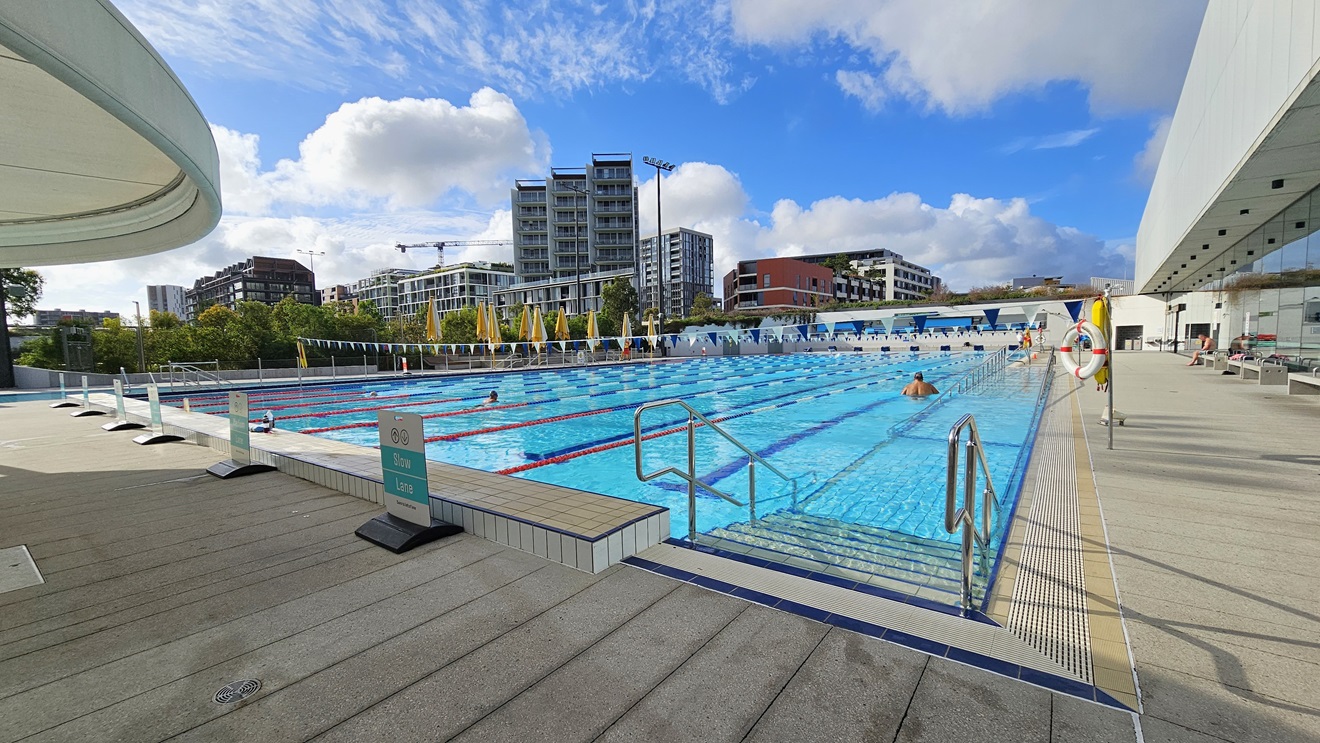 The 50m Pool is equipped with stepped entries with handrails. Safety banners are arranged on the pool deck on the left. On the right, a hoist is positioned near the pool's overflow grid. Colorful floating ropes and a string of triangle banners run across the pool. Bench chairs are arranged on the right near the glass wall. To the left, a section of the circular shade structure is also visible. Further, tall buildings are also visible.