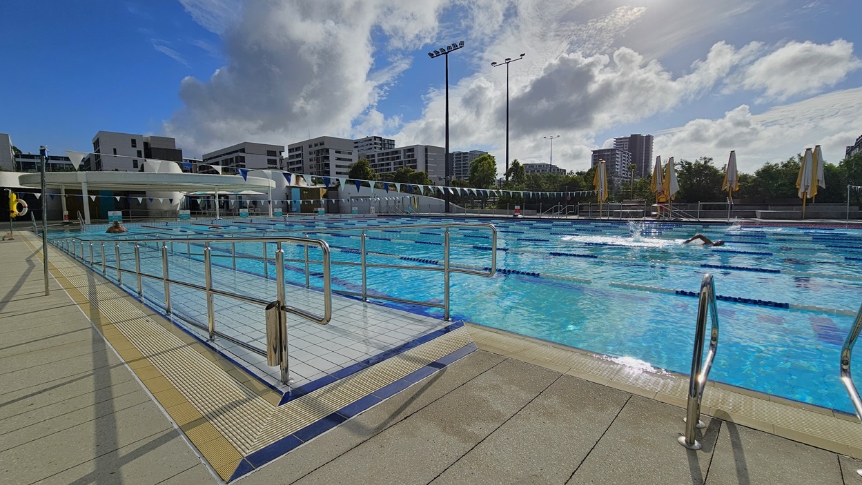 The 50m Pool is equipped with a ramp and stepped entries with handrails. Safety banners and a hoist are positioned on the far left near the pool overflow grid. Colorful floating ropes and a string of triangle banners run across the pool. On the right, folded yellow umbrella shades. Further, tall floodlights and buildings are also visible.