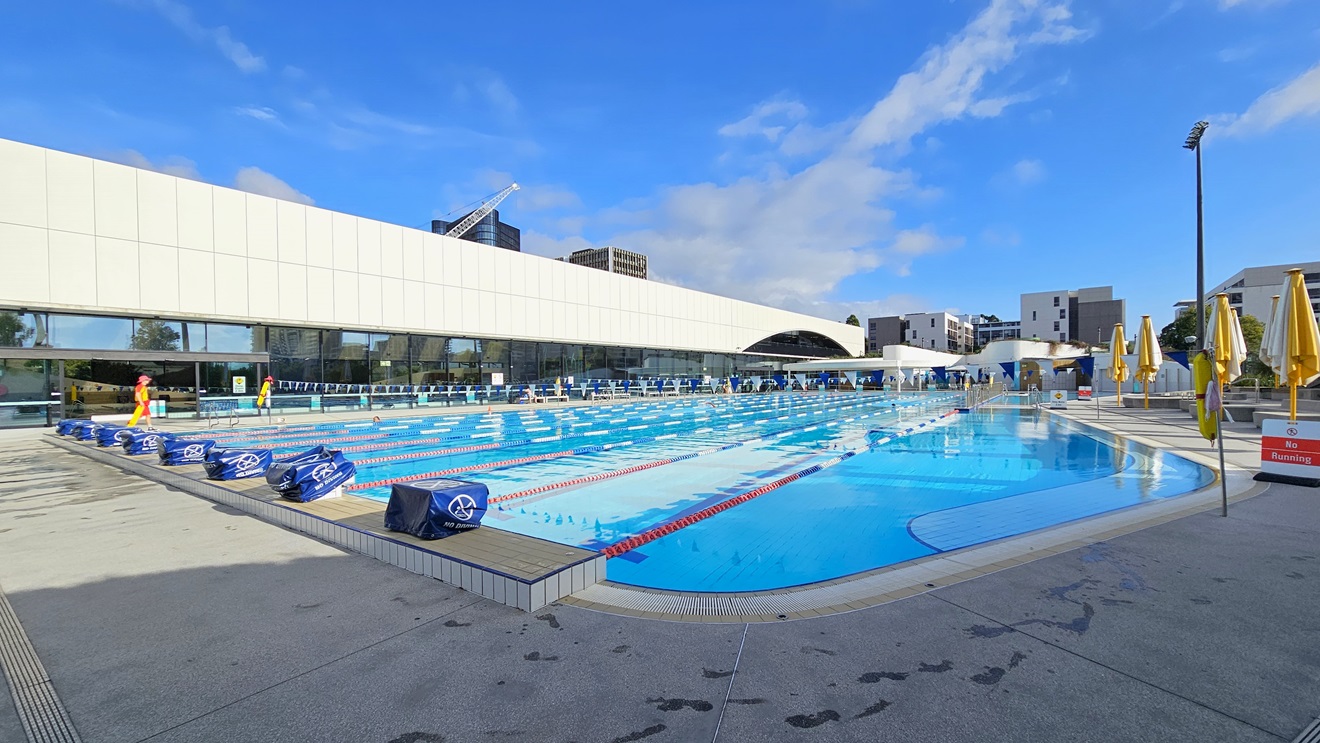 To the left is the facility building equipped with a white-tiled facade and glass walls. The 50m Pool is equipped with diving boards with covers. Safety banners and a hoist are positioned on the right near the pool overflow grid, along with folded umbrella shades. Colorful floating ropes and a string of triangle banners run across the pool. On the right, folded yellow umbrella shades. Tall floodlights and buildings are also visible.