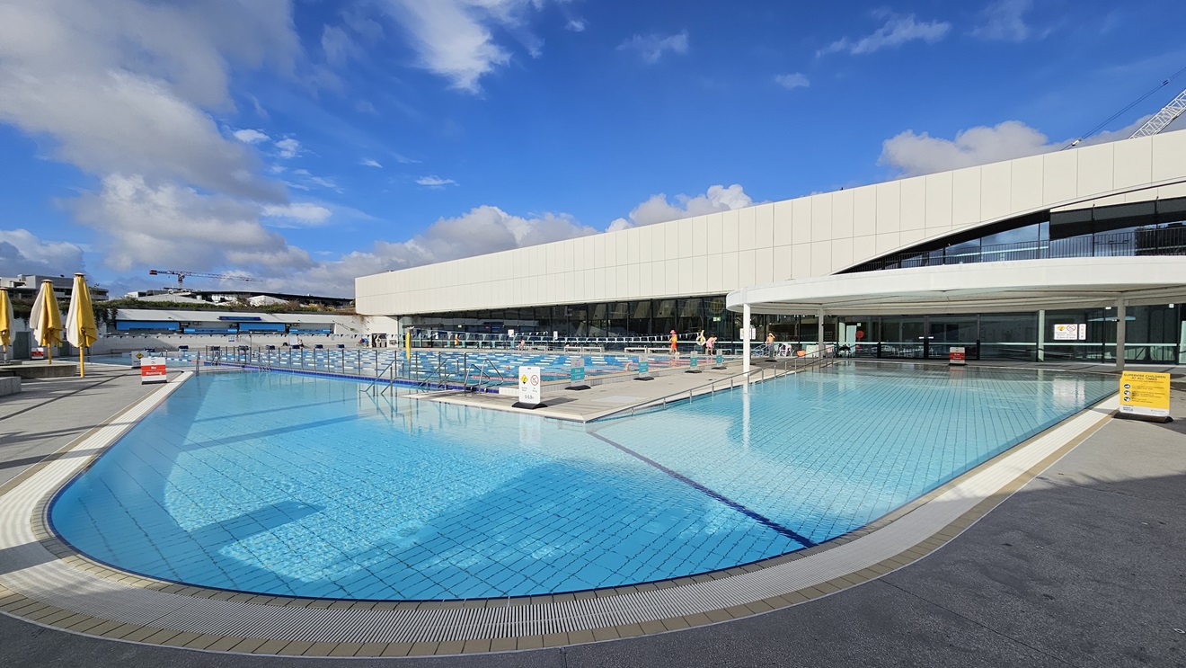 The Leisure Pool features stairs with a ramp entry and is surrounded by an overflow grid. On the right is a circular shade structure that offers shading to the Leisure Pool. The facility building is also adjacent. Adjacent to it is the 50m Pool equipped with floating ropes and triangle banners. In between these two pools is a pool deck equipped with safety banners. On the left, folded umbrella shades are also visible.