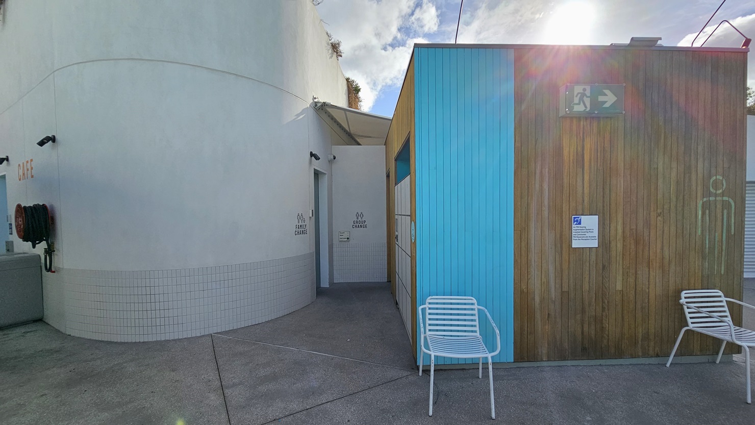 To the right is a Cabana-style Male and Female Change Room featuring a wooden design with a blue accent exterior. Signage is fixed to the wall. White chairs are positioned. To the left is a family change room featuring a concrete grey wall with small tiles accent. A fire hose reel is positioned on the left.