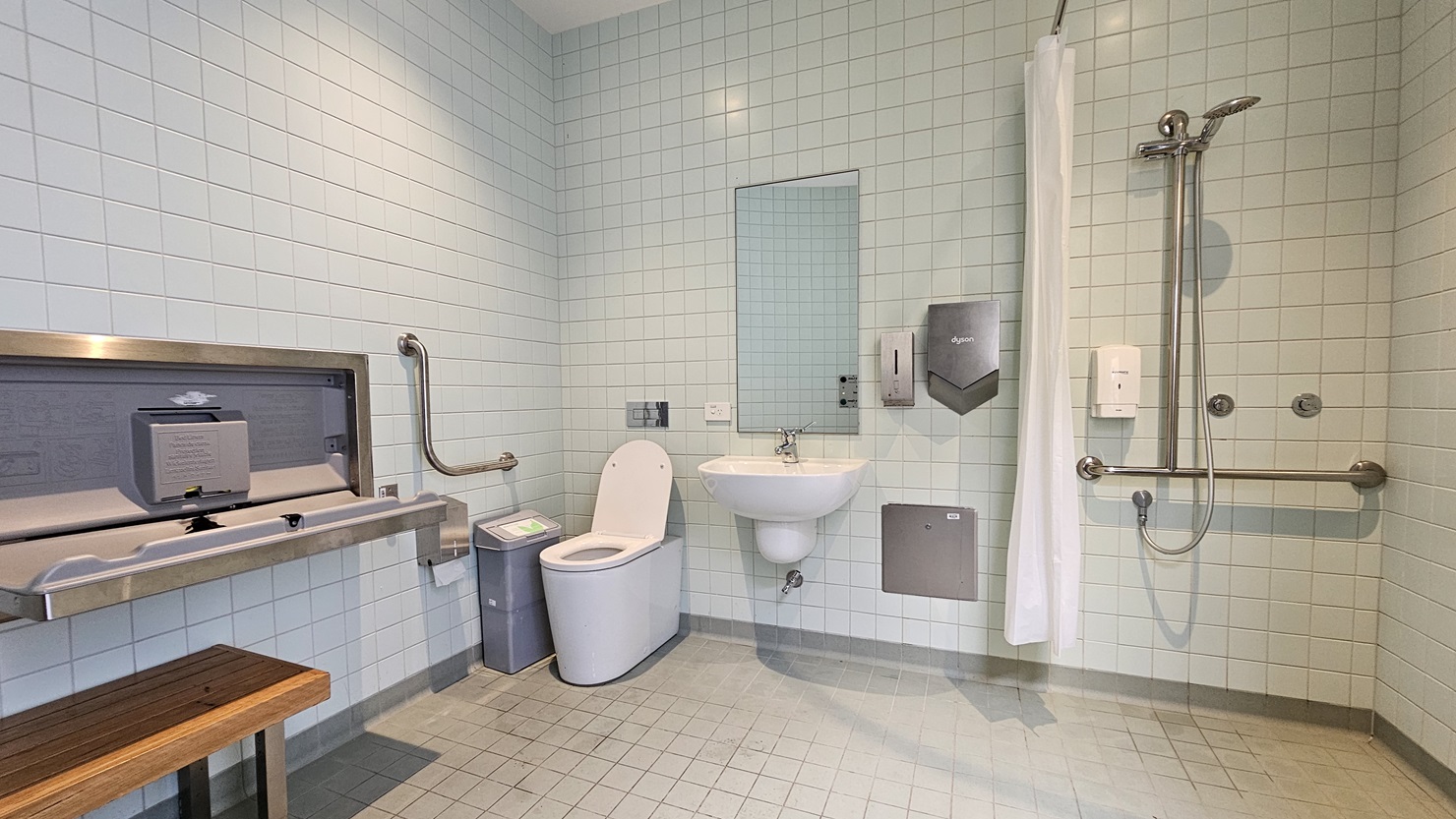 To the right is a shower equipped with handrails and a shower curtain. Next to it is the toilet bowl with handrails. A mirror, soap, and tissue dispenser are affixed to the wall. A rubbish bin and toilet brush are placed nearby. To the left is a wooden bench for seating and a foldable change table. The walls are covered with white tiles from floor to ceiling. The ceiling is white with LED lights, and the floor has white tiles.