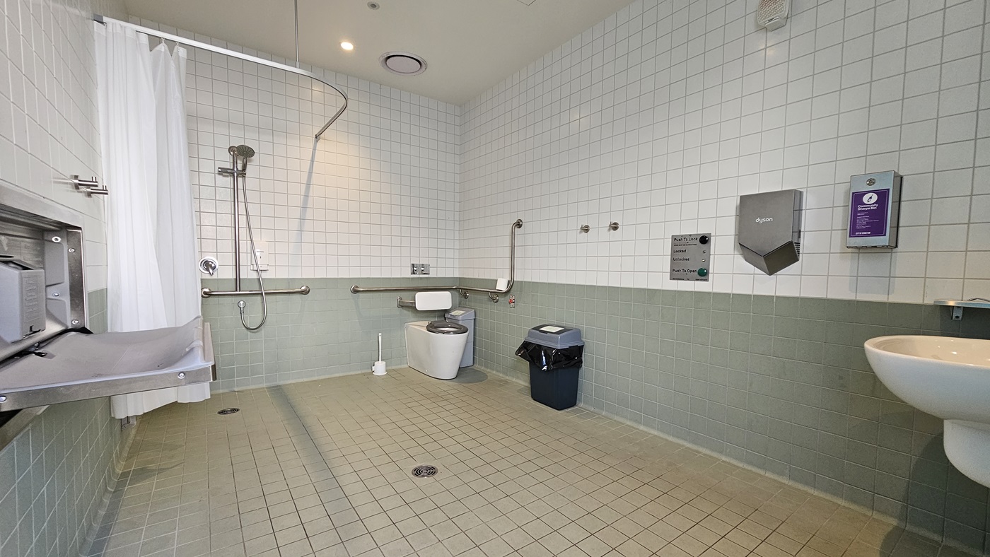 To the left is a shower equipped with handrails and a shower curtain. Next to it is the toilet bowl with hand grabs. A rubbish bin and toilet brush are placed nearby. To the left, a foldable change table. A mirror, soap, and tissue dispenser are affixed to the right wall, along with emergency buttons. The walls are covered with white tiles from floor to ceiling. The ceiling is white with LED lights, and the floor has white tiles.