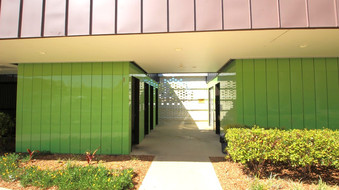 A view of the bathrooms' exterior. There is an aisle between two separated divisions of the lime-green coated painted bathroom walls facing each other mounted with overhead bracings and several standard doors. A view of the bathrooms' exterior. There is an aisle between two separated divisions of the lime-green coated painted bathroom walls facing each other mounted with overhead bracings and several standard doors.