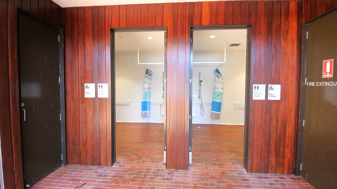 Entrance leading to the bathrooms. Signage can be found on both bathroom doors, conveying that these facilities offer unisex toilets with showers, baby-changing facilities, as well as separate male and female restrooms and showers. Furthermore, there is a rubbish bin located to the right of the restroom entrance. Adjacent to the bathroom, on the right side, there is also a fire extinguisher logo visible. Entrance leading to the bathrooms. Signage can be found on both bathroom doors, conveying that these facilities offer unisex toilets with showers, baby-changing facilities, as well as separate male and female restrooms and showers. Furthermore, there is a rubbish bin located to the right of the restroom entrance. Adjacent to the bathroom, on the right side, there is also a fire extinguisher logo visible.