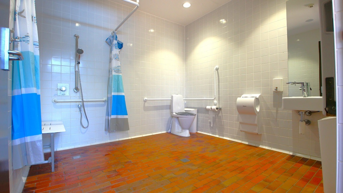 Another view of a bathroom featuring the shower to the left with a blue shaded shower curtain whilst to the right is the toilet. Further to the right is the hand basin and hand dryer. Another view of a bathroom featuring the shower to the left with a blue shaded shower curtain whilst to the right is the toilet. Further to the right is the hand basin and hand dryer.