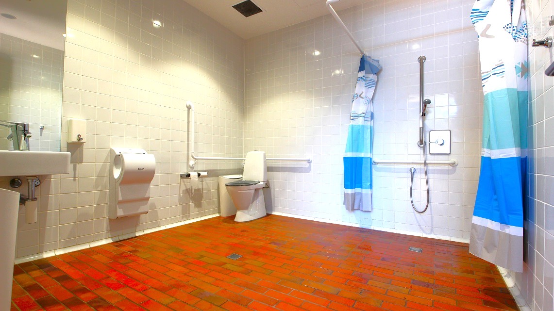 Square ceramic, light-colored tiles cover the bathroom walls. Patterned blue and white curtains draped over the rails served as partitions for privacy. Bright light fills the space overhead through bathroom ceiling lighting. On the other corner is a vanity area of the sink and mirror adjacently positioned to the showerhead and towel racks. Square ceramic, light-colored tiles cover the bathroom walls. Patterned blue and white curtains draped over the rails served as partitions for privacy. Bright light fills the space overhead through bathroom ceiling lighting. On the other corner is a vanity area of the sink and mirror adjacently positioned to the showerhead and towel racks.