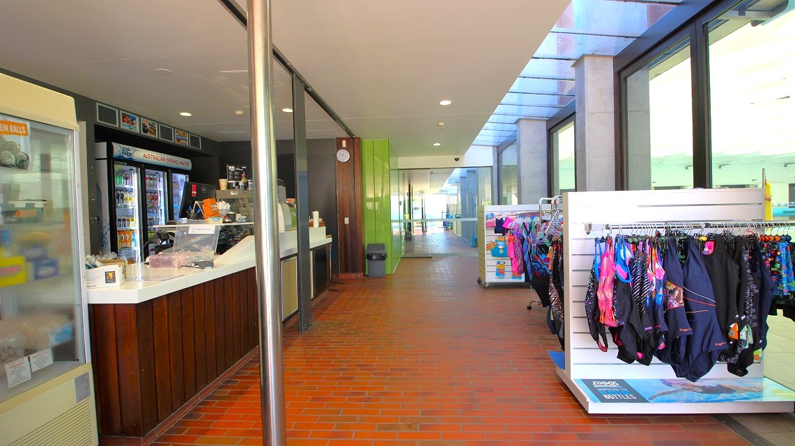 Pathway adjacent to the reception area. To the right, an array of swimwear is elegantly showcased, drawing attention away from the reception area. Inside the reception space, beverages are prominently on display, while framed artwork adorns the walls, adding to the aesthetic appeal and suggesting a focus on both fashion and refreshment. Pathway adjacent to the reception area. To the right, an array of swimwear is elegantly showcased, drawing attention away from the reception area. Inside the reception space, beverages are prominently on display, while framed artwork adorns the walls, adding to the aesthetic appeal and suggesting a focus on both fashion and refreshment.