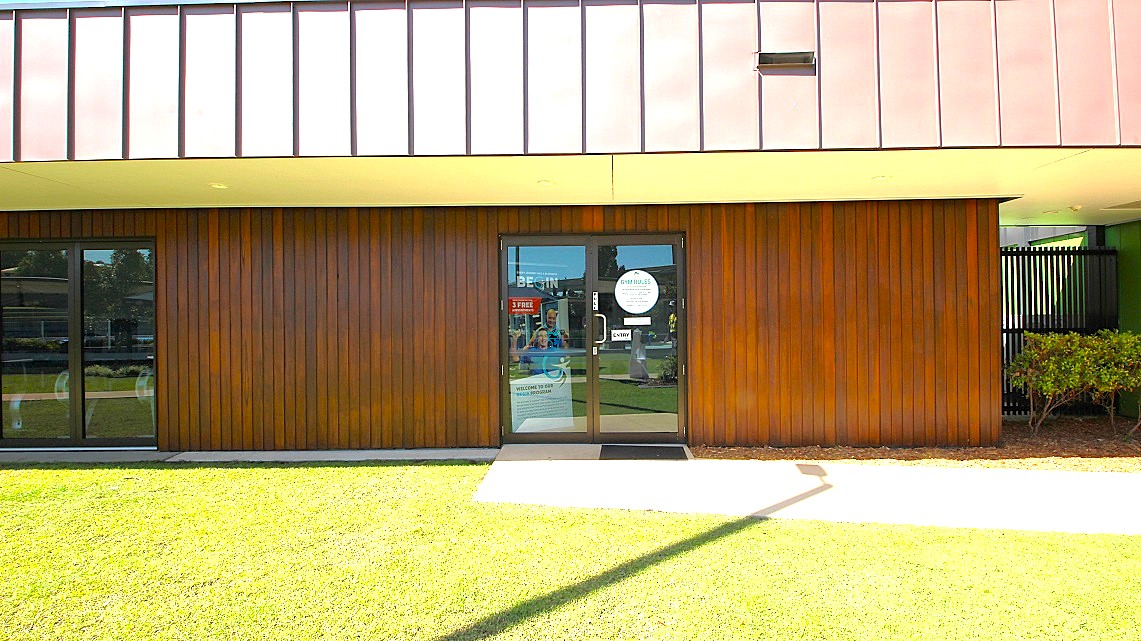 Outdoor look of the Gymnasium’s main entrance. The exterior space is adorned with well-manicured lawns bordering the walkways leading to the gymnasium. The manual glass doors are notably adorned with multiple informative signs. On the right corner, just behind the decorative elements, there stands a tall black fence. Outdoor look of the Gymnasium’s main entrance. The exterior space is adorned with well-manicured lawns bordering the walkways leading to the gymnasium. The manual glass doors are notably adorned with multiple informative signs. On the right corner, just behind the decorative elements, there stands a tall black fence.