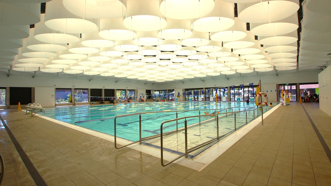 A closer look at the ramp access of the pool. The indoor pool is well-lit, with an array of ceiling lights creating a bright atmosphere. Inside the pool, blue-colored lane ropes and flags are positioned at both ends. These lane ropes separate swim lanes, and the flags help swimmers gauge their position, contributing to a safe and organized swimming environment. A closer look at the ramp access of the pool. The indoor pool is well-lit, with an array of ceiling lights creating a bright atmosphere. Inside the pool, blue-colored lane ropes and flags are positioned at both ends. These lane ropes separate swim lanes, and the flags help swimmers gauge their position, contributing to a safe and organized swimming environment.