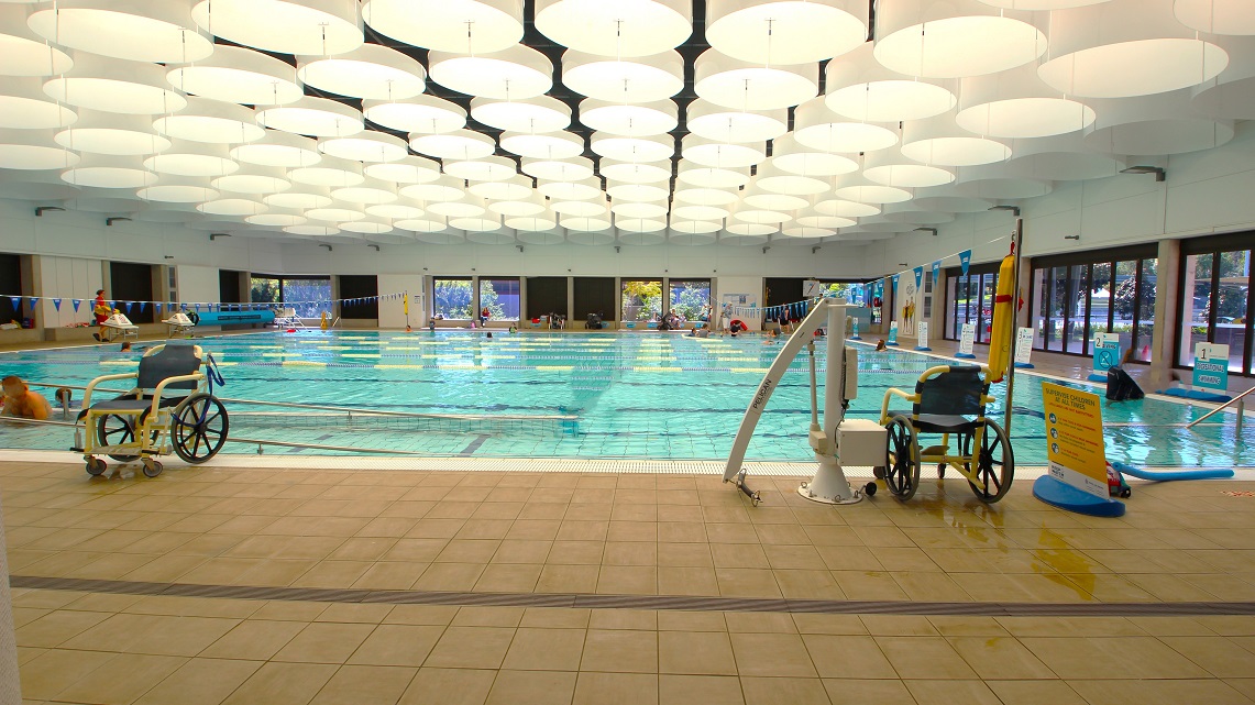 Inside the Indoor Heated Pool. The indoor pool at Gympie ARC spans 25 meters and is entirely enclosed. It maintains a comfortable temperature range between 28°C and 29.5°C and offers access through both ramps and steps. Natural light streams in through glass windows on two sides. Additionally, you can spot a couple of aquatic wheelchairs and a pool hoist, with informative signage positioned next to one of the aquatic wheelchairs. Inside the Indoor Heated Pool. The indoor pool at Gympie ARC spans 25 meters and is entirely enclosed. It maintains a comfortable temperature range between 28°C and 29.5°C and offers access through both ramps and steps. Natural light streams in through glass windows on two sides. Additionally, you can spot a couple of aquatic wheelchairs and a pool hoist, with informative signage positioned next to one of the aquatic wheelchairs.