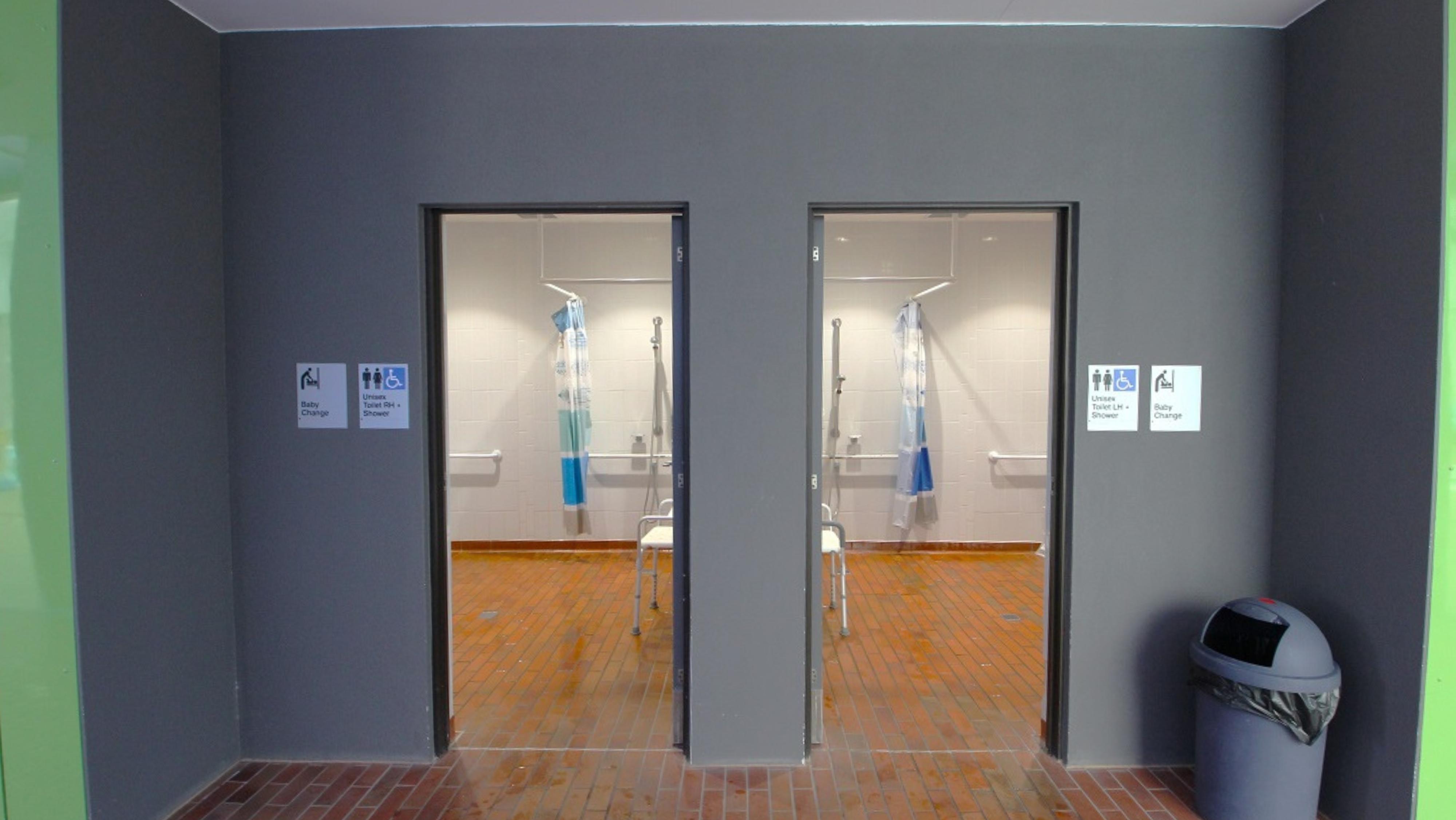 Entrance leading to the accessible bathrooms. Signage is present on both restroom doors, indicating that these facilities provide unisex toilets equipped with showers, baby changing amenities, and male and female restrooms and showers. Additionally, there's a rubbish bin positioned to the right of the restroom entrance. Entrance leading to the accessible bathrooms. Signage is present on both restroom doors, indicating that these facilities provide unisex toilets equipped with showers, baby changing amenities, and male and female restrooms and showers. Additionally, there's a rubbish bin positioned to the right of the restroom entrance.