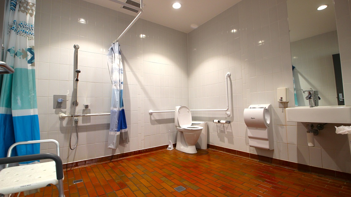 Inside the unisex bathroom. The bathroom distinguishes itself through the use of unique brick floor tiles, making it noticeably different from the surrounding area. Additionally, the toilet, sink, showerhead, toilet paper holder, and towel racks are all placed in a well-organized and spacious manner. Moreover, the bathroom is brightly illuminated with fluorescent lighting, ensuring excellent visibility. Inside the unisex bathroom. The bathroom distinguishes itself through the use of unique brick floor tiles, making it noticeably different from the surrounding area. Additionally, the toilet, sink, showerhead, toilet paper holder, and towel racks are all placed in a well-organized and spacious manner. Moreover, the bathroom is brightly illuminated with fluorescent lighting, ensuring excellent visibility.