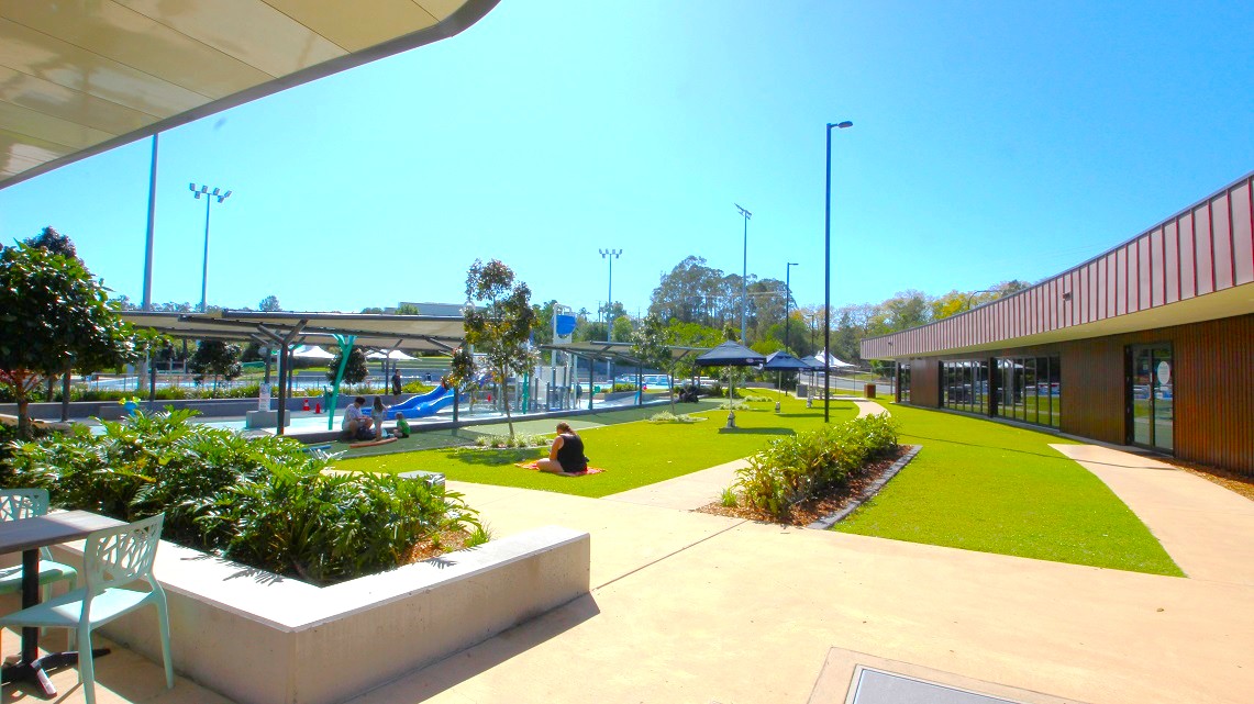 On the left, there is a seating area with a table and chairs next to a planter filled with greenery. In the centre, a person is sitting on a grassy area, surrounded by small trees and plants. To the right, a modern building with large windows runs along the edge of the scene. In the background, there is a playground with slides and a covered area, along with tall light poles and more trees. The sky is clear and blue. On the left, there is a seating area with a table and chairs next to a planter filled with greenery. In the centre, a person is sitting on a grassy area, surrounded by small trees and plants. To the right, a modern building with large windows runs along the edge of the scene. In the background, there is a playground with slides and a covered area, along with tall light poles and more trees. The sky is clear and blue.