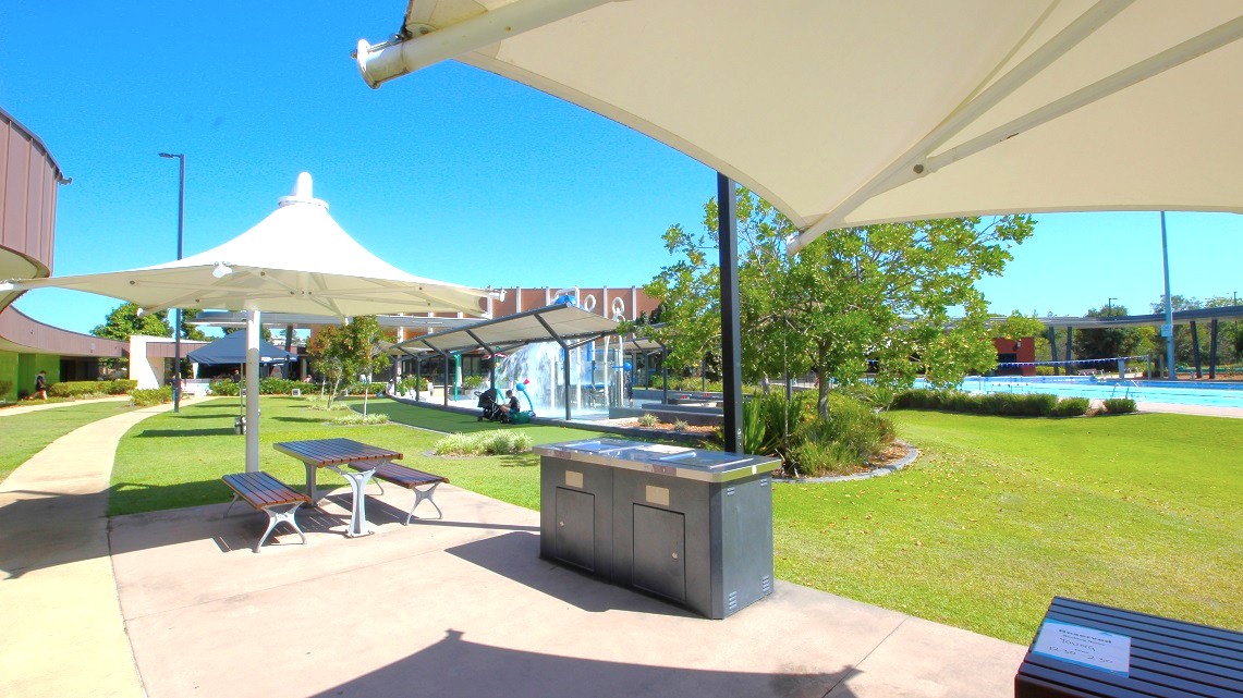 A park area with a paved walkway and green grass. On the left, there are two picnic tables under large white shade structures. In the centre, a barbecue grill is visible. To the right, there are trees and a glimpse of a swimming pool. In the background, a building with a glass facade and more greenery can be seen. The sky is clear and blue. A park area with a paved walkway and green grass. On the left, there are two picnic tables under large white shade structures. In the centre, a barbecue grill is visible. To the right, there are trees and a glimpse of a swimming pool. In the background, a building with a glass facade and more greenery can be seen. The sky is clear and blue.