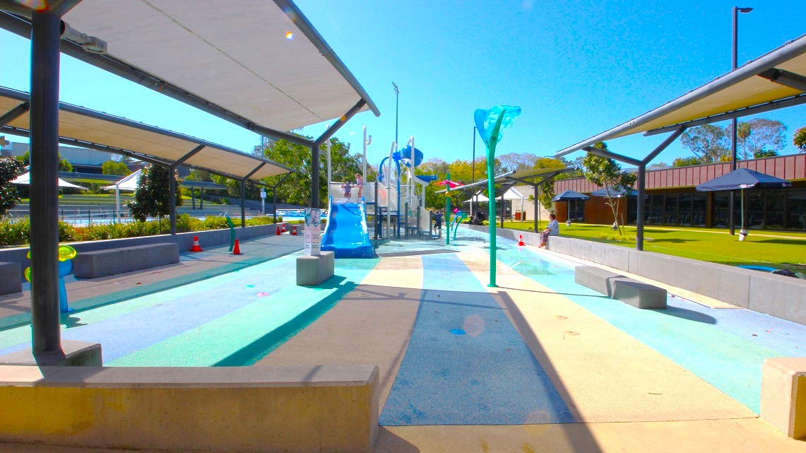A water play area with a central pathway featuring a blue slide and various water features. On both sides, there are shaded structures with benches underneath. The ground is painted in shades of blue and beige, resembling water. In the background, there are trees and a building with a dark roof. The area is open and sunny, with a clear blue sky above. A water play area with a central pathway featuring a blue slide and various water features. On both sides, there are shaded structures with benches underneath. The ground is painted in shades of blue and beige, resembling water. In the background, there are trees and a building with a dark roof. The area is open and sunny, with a clear blue sky above.