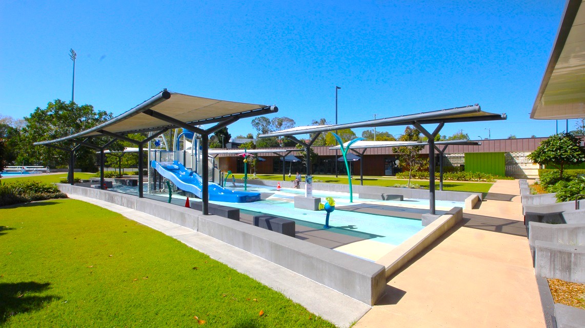 A children's water play area with a blue slide in the centre, surrounded by shallow pools. Overhead, there are shade structures with metal frames. The ground is a mix of concrete paths and green grass. In the background, there are trees and a building with a brown roof. A few colourful water play features are scattered around the area. A children's water play area with a blue slide in the centre, surrounded by shallow pools. Overhead, there are shade structures with metal frames. The ground is a mix of concrete paths and green grass. In the background, there are trees and a building with a brown roof. A few colourful water play features are scattered around the area.