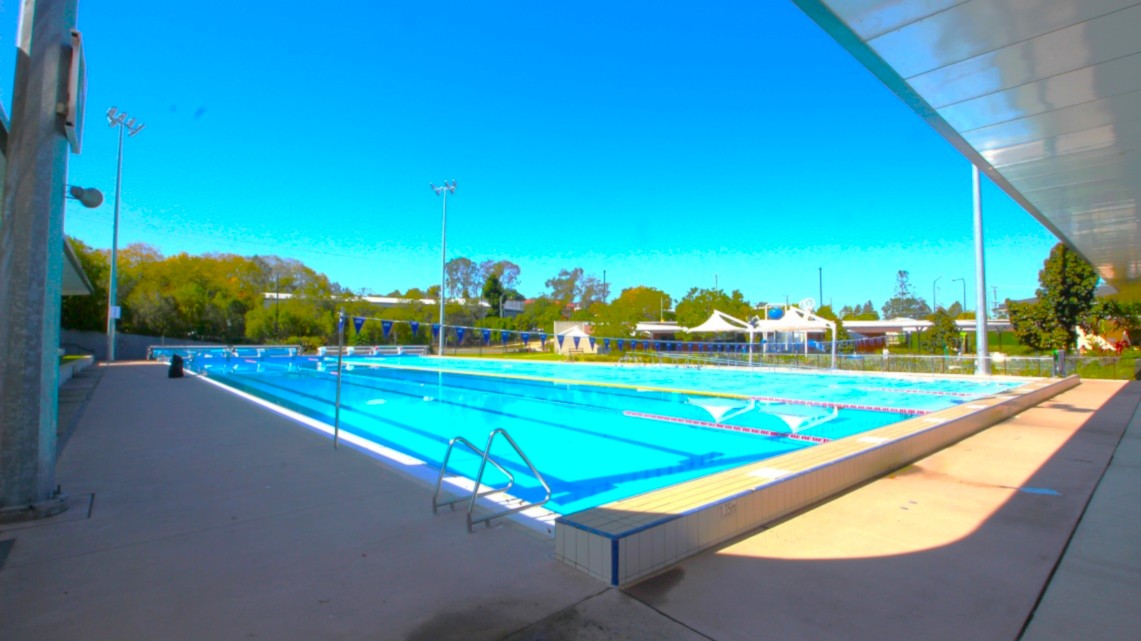 An outdoor swimming pool with clear blue water, surrounded by a concrete deck. On the left, there are tall floodlights and a small building. The right side features a covered walkway with a metal roof. In the background, there are trees and several white tents or canopies. The pool has lane dividers, and there are metal ladders for entry. The area is well-lit, and the sky is clear. An outdoor swimming pool with clear blue water, surrounded by a concrete deck. On the left, there are tall floodlights and a small building. The right side features a covered walkway with a metal roof. In the background, there are trees and several white tents or canopies. The pool has lane dividers, and there are metal ladders for entry. The area is well-lit, and the sky is clear.